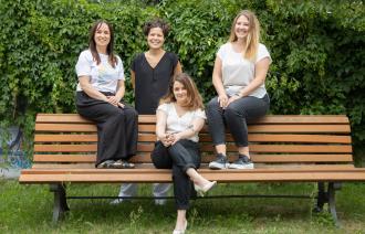 Die Beauftragten des Büros für Chacengleichheit und Vielfalt sitzen auf einer braunen Bank,  im Hintergrund ist eine grüne Hecke zu sehen (von links nach rechts: Stefanie Buhr, Dr. Tina Denninger, Claudia Sprengel, Dr. Amanda Palenberg)