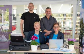 Zwei Frauen sitzen an einem Arbeitsplatz, der über einen Laptop und einen Druck verfügt. Hinter den beiden Frauen stehen zwei Männer. Alle schauen in die Kamera.