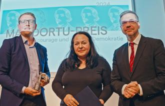 Drei Personen stehen auf der Bühne der Preisverleihung, darunter die Preisträgerin Katharina König-Preuss