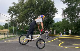 Eine Jugendliche steht auf einem BMX-Rad und fährt über die asphaltierte Berg- und Tal-Bahn, die Pumptrack genannt wird.