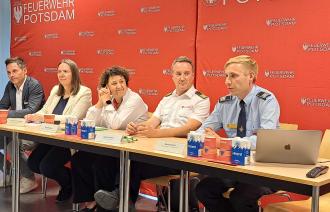 Fünf Menschen sitzen vor einer Pressewand der Feuerwehr Potsdam bei einer Pressekonferenz.
