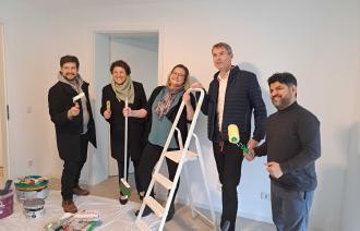 Fünf Personen stehen in einem Raum und halten Utensilien für eine Wohnungsrenovierung in der Hand.