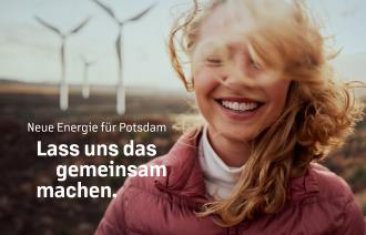 Eine junge Frau steht lachend und mit von Wind verwehtem Haar vor einer Landschaft, in der Windräder gebaut sind.
