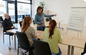 Eine Frau steht vor einem Flipchart und erklärt etwas. Ihr zugewandt sitzen drei Personen an Tischen, die ihr zuhören.