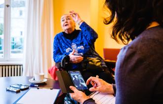 Das Bild zeigt eine Frau, die auf einem Handy im Internet surft, sowie eine ältere sitzende Dame, die etwas erzählt.