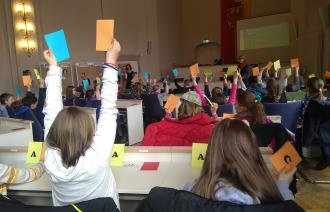 Viele Kinder sitzen in einem großen Saal und halten bunte Abstimmungskarten in die Luft. Sie nehmen an einer Mitmachveranstaltung teil, bei der Fragen beantwortet werden