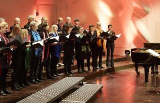 23 Mitglieder des Claudius-Ensembles singen auf der Bühne der Nikolaisaal; ein Flügel und ein Dirigierpult sind ebenfalls zu sehen.