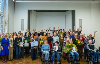 „Großes Gruppenfoto der Preisträgerinnen und Preisträger des Potsdamer Inklusionspreises 2025. Menschen mit und ohne Behinderung stehen und sitzen gemeinsam im Saal des Potsdam Museums, viele halten Blumen und Urkunden, alle lächeln und jubeln.“