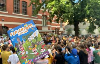 Eine Person hält mit der rechten Hand den Ferienpass für 2024. Im Hintergrund sieht man viele sitzende Personen.