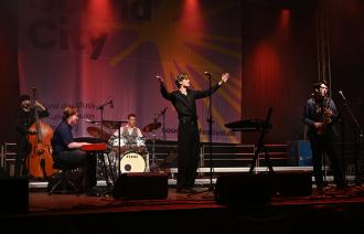  Eine Pop-Jazzband mit 5 jungen Männern performed auf professionell beleuchteter Bühne - der Sänger breitet seine Arme aus