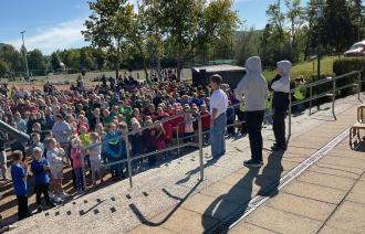 Wir schauen von hinten auf drei Jugendliche, die einen Song darbieten. Gemeinsam mit ihnen sehen hunderte Kinder und Jugendliche als Publikum auf einem Sportplatz