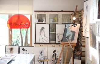 Blick in ein Kunstatelier, in dem sich viele Bilder, eine rote Lampe, ein weißer Schrank, eine Staffelei und mehrere Bücher befinden