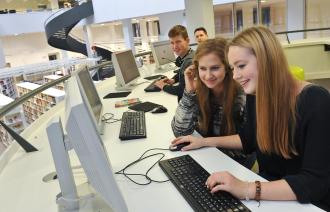 Zwei Jugendliche sitzen vor einem Computer - im Hintergrund sitzen zwei weitere Jugendliche. 