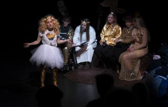 Im Vordergrund steht eine geschminkte Person mit blonder Perücke, weißem Rock und goldenen Stiefeln. Im Hintergrund sitzen weitere fünf kostümierte Personen im Halbkreis.