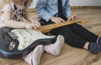 Zwei Kinder sitzen auf dem Boden mit einer E-Gitarre auf dem Schoß.