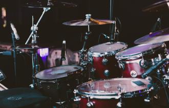 Wir schauen von schräg hinten in ein spannende beleuchtetes Drumset. Es ist mikrofoniert und steht in einem Tonstudio oder einer Konzertbühne.