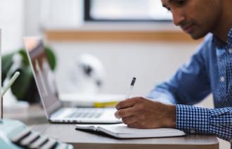 Mann (im Profil) sitzt an einem Tisch und macht sich Notizen. Ein aufgeklappter Laptop steht im Hintergrund. Es könnte sich um einen Co-Working-Space handeln. Der Mann trägt ein weiß-blau-kariertes Hemd. Er hat eine dunkle Hautfarbe.