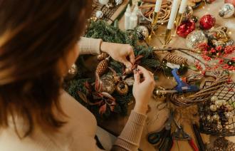 Eine Frau beim Basteln. Sie verziert einen Adventskranz. Blick auf die Hände, von hinten oben. Weihnachtliche Bastelmaterialien liegen umher.