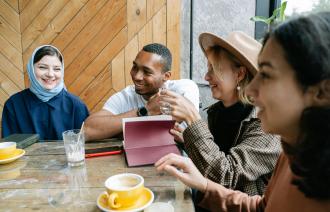 Vier Personen sitzen zusammen an einem Tisch. Sie lachen vor ihnen stehen Tassen und ein Glas. Eine Person schaut aiuf ein Tablet. Eine Frau trägt ein Kopftuch, auch ist ein Mann mit dunkler Hautfarbe dabei. Das Bild vermittelt Diversität und Gemeinschaft.