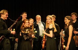 Einige junge klassische Musikerinnen und Musiker freuen sich über einen gelungenen Auftritt und posieren mit ihren Instrumenten. In ihrer Mitte der Leiter der Gruppe aus Opole namens Hubert Prochota. Er ist weißhaarig und hat einen weissen Bart. Auch er freut sich sichtlich!