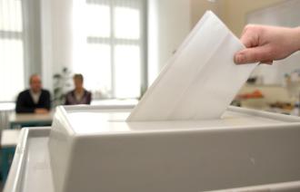 Wahlurne im Vordergrund mit Hand die Wahlschein einsteckt und zwei Personen unscharf im Hinterrund