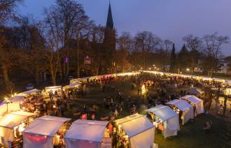Beleuchteter Weihnachtsmarkt am Abend vor einer Kirche, mit vielen Besuchern, weißen Zelten und kahlen Bäumen., Foto: André Stiebitz, Lizenz: PMSG