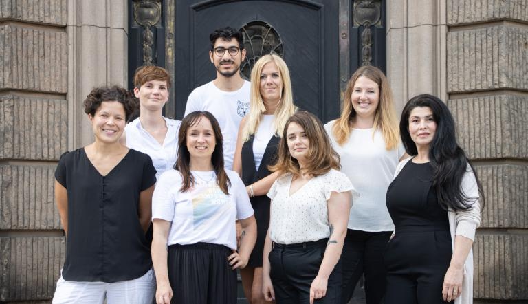 Die Teammitglieder des Büros für Chancengleichheit und Vielfalt der Landeshauptstadt Potsdam stehen auf einer Treppe, hinter ihnen sieht man mittig eine Eingangstür und links und rechts daneben Hauswände (erste Reihe von links nach rechts: Dr. Tina Denninger, Stefanie Buhr, Claudia Sprengel, Sindy Valverde; zweite Reihe von links nach rechts: Ina Bellmann, Susanne Plath, Dr. Amanda Palenberg; dritte Reihe: Kareem Khader)