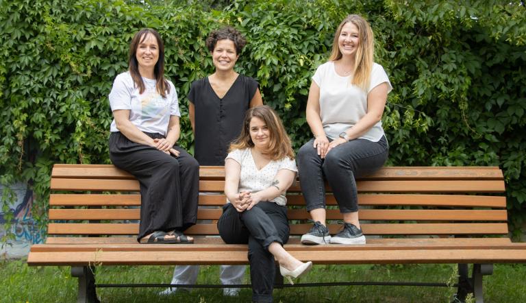 Die Beauftragten des Büros für Chacengleichheit und Vielfalt sitzen auf einer braunen Bank,  im Hintergrund ist eine grüne Hecke zu sehen (von links nach rechts: Stefanie Buhr, Dr. Tina Denninger, Claudia Sprengel, Dr. Amanda Palenberg)
