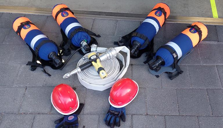 Die neuen Übungs-Atemschutzgeräte samt Ausrüstung für die Jugendfeuerwehr. Vier neue Atemschutzgeräte für die Jugendfeuerwehr liegen auf dem Boden, danaben rote Helme, Handschuhe und ein Feuerwehrschlauch mit Spritze.
