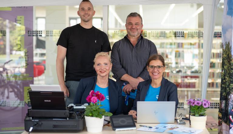 Zwei Frauen sitzen an einem Arbeitsplatz, der über einen Laptop und einen Druck verfügt. Hinter den beiden Frauen stehen zwei Männer. Alle schauen in die Kamera.