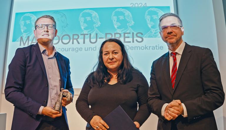 Drei Personen stehen auf der Bühne der Preisverleihung, darunter die Preisträgerin Katharina König-Preuss