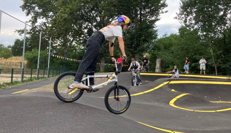 Eine Jugendliche steht auf einem BMX-Rad und fährt über die asphaltierte Berg- und Tal-Bahn, die Pumptrack genannt wird.