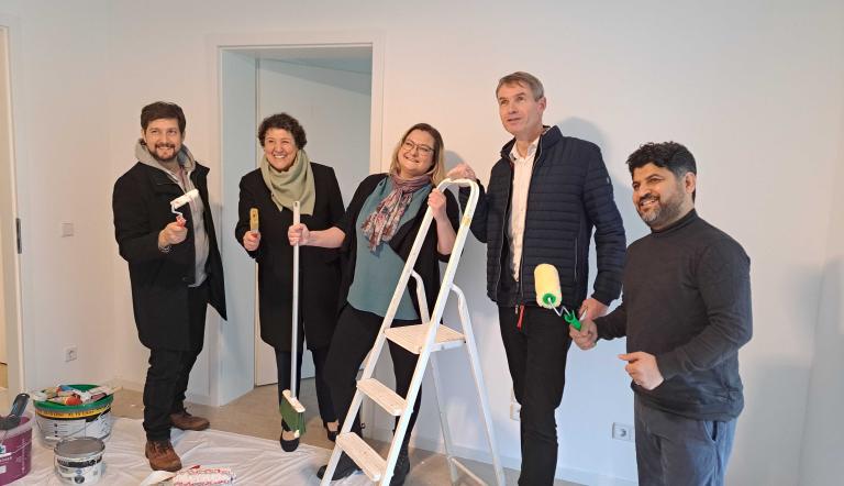 Fünf Personen stehen in einem Raum und halten Utensilien für eine Wohnungsrenovierung in der Hand.