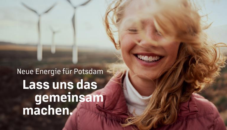 Eine junge Frau steht lachend und mit von Wind verwehtem Haar vor einer Landschaft, in der Windräder gebaut sind.