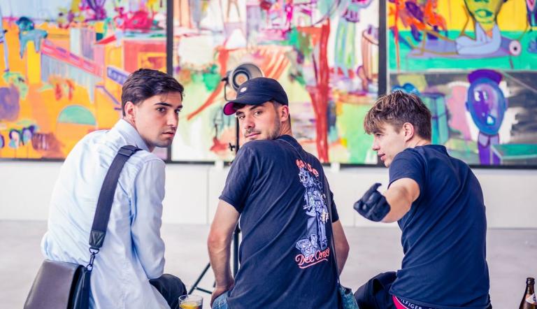 3 junge Männer sitzen auf einer Bank vor einer bunt bemalten Wand