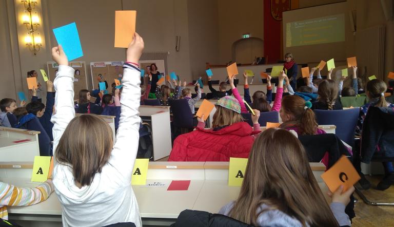Viele Kinder sitzen in einem großen Saal und halten bunte Abstimmungskarten in die Luft. Sie nehmen an einer Mitmachveranstaltung teil, bei der Fragen beantwortet werden