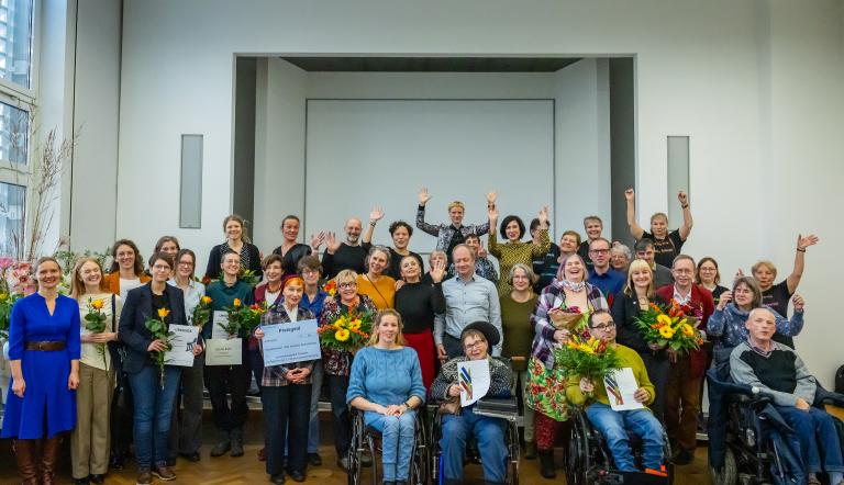 „Großes Gruppenfoto der Preisträgerinnen und Preisträger des Potsdamer Inklusionspreises 2025. Menschen mit und ohne Behinderung stehen und sitzen gemeinsam im Saal des Potsdam Museums, viele halten Blumen und Urkunden, alle lächeln und jubeln.“