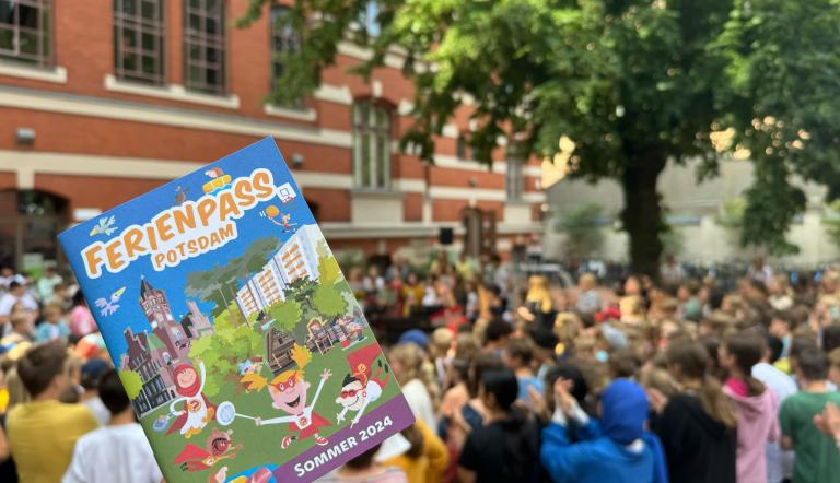 Eine Person hält mit der rechten Hand den Ferienpass für 2024. Im Hintergrund sieht man viele sitzende Personen.