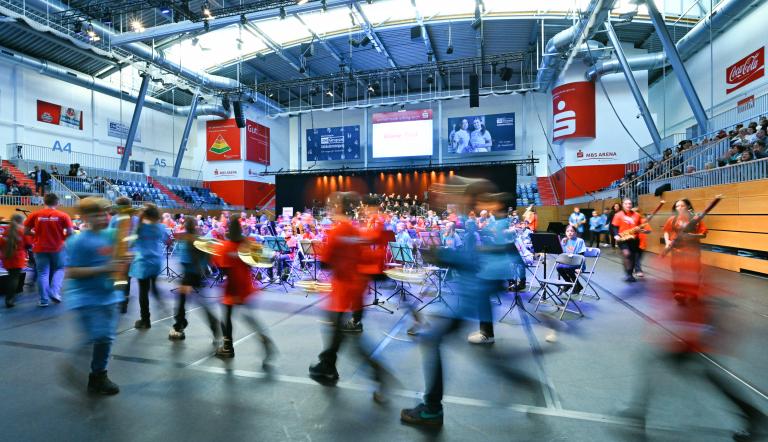 Klasse : Musik - Einzug in die MBS-Arena Viele Kinder mit Musikinstrumenten laufen so schnell in die große MBS Arena, dass sie nur unscharf zu erkennen sind