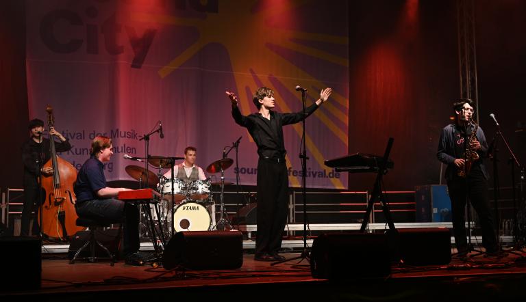  Eine Pop-Jazzband mit 5 jungen Männern performed auf professionell beleuchteter Bühne - der Sänger breitet seine Arme aus