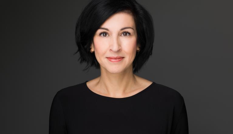 Noosha Aubel hat schwarze, schulterlange Haare. Sie trägt Lippenstift und ein schwarzes Oberteil. Sie wirkt sympathisch.