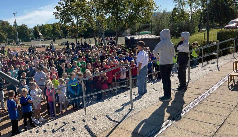 Wir schauen von hinten auf drei Jugendliche, die einen Song darbieten. Gemeinsam mit ihnen sehen hunderte Kinder und Jugendliche als Publikum auf einem Sportplatz