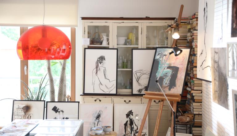 Blick in ein Kunstatelier, in dem sich viele Bilder, eine rote Lampe, ein weißer Schrank, eine Staffelei und mehrere Bücher befinden