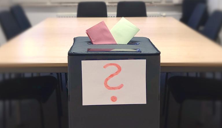 In einer Wahlurne stecken zwei bunte Zettel und an der Wahlurne hängt ein Zettel mit einem Fragezeichen.