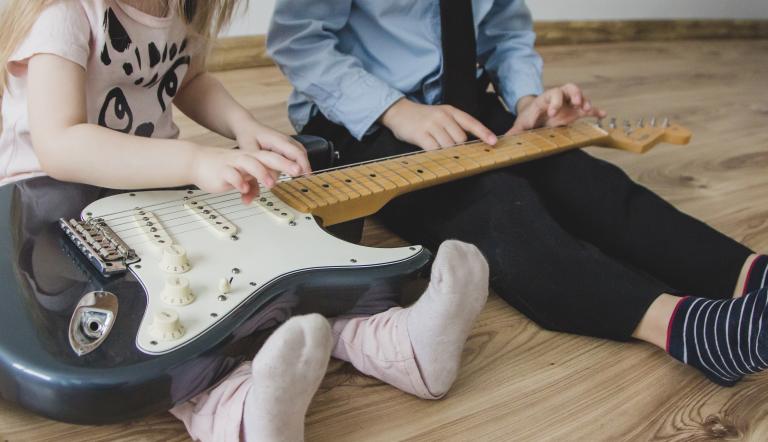 Zwei Kinder sitzen auf dem Boden mit einer E-Gitarre auf dem Schoß.