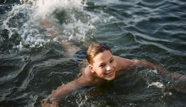 Eine Junge schwimmt in einem See und schaut lächelnd in die Kamera..