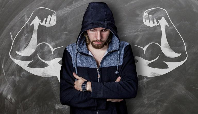 Ein Mann träumt davon, stark zu sein. Er ist niedergeschlagen, trägt einen Kapuzenpulli. Hinter ihm sieht man starke, muskelbepackte Arme, die ihm wie Flügel zu wachsen scheinen. Eine Kreidezeichnung auf grauem Grund im Bildhintergrund.