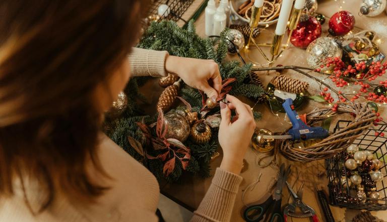 Eine Frau beim Basteln. Sie verziert einen Adventskranz. Blick auf die Hände, von hinten oben. Weihnachtliche Bastelmaterialien liegen umher.