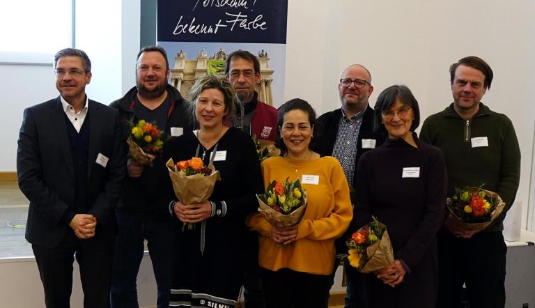 Das Foto zeigt die sieben Mitglieder des Lenkungsgremiums neben dem Potsdamer Oberbürgermeister. Die Gruppe steht vor einem "Potsdam! bekennt Farbe"-Plakat.