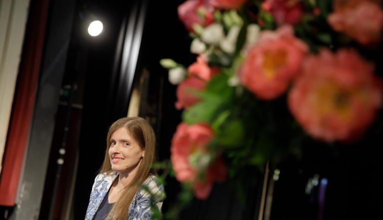 Links sieht man Adina Hermann, rechts im Bild sieht man verschwommen weiße und pfirschfarbene Blumen